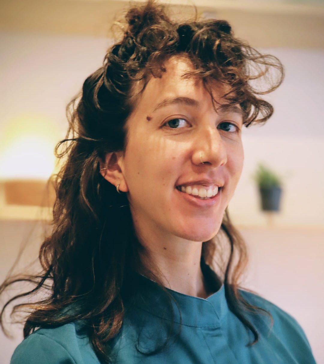 Femme souriante avec des cheveux frisés, portant un haut bleu, dans un intérieur lumineux. Photo présentant la praticienne de massage