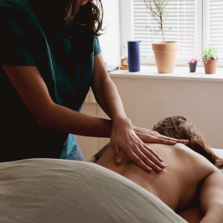Femme recevant un massage, avec une praticienne en arrière-plan. Plantes en pot visibles.